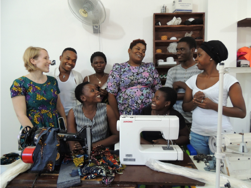 Kitenge founder Sian standing with our accessories tailor better and her team of talented tailors inside their brand new larger workshop in Tanzania in front of a table of sewing machines