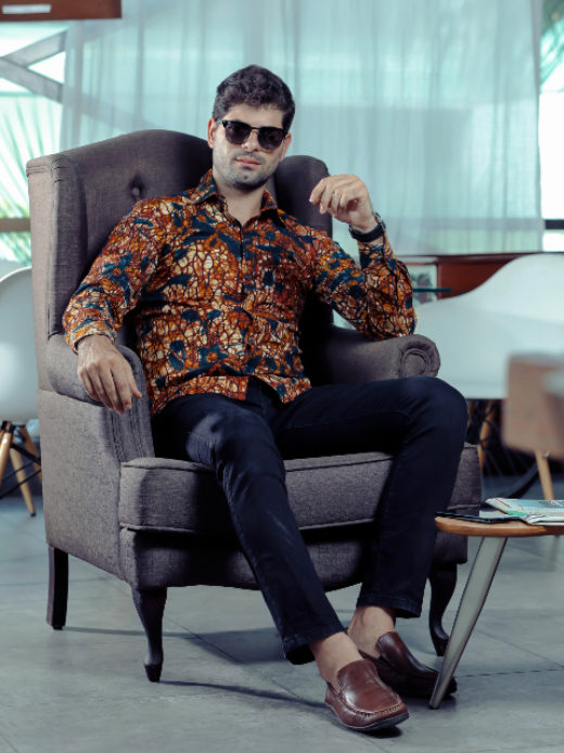Men's gold floral custom-made African print long sleeve shirt model sat in arm chair wearing sunglasses