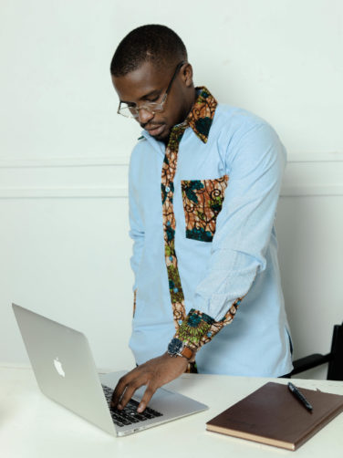 Men's custom-made plain light blue long sleeve shirt with optional African wax print fabric contrasts model wearing using laptop